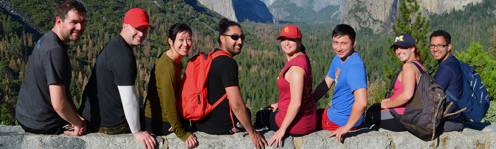 Group of people sit on rock ledge with foliage and mountains in the background.