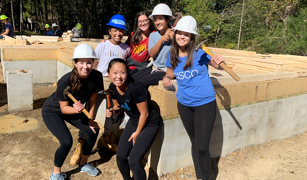 Six people wearing safety helmets pose together smiling on a construction site.